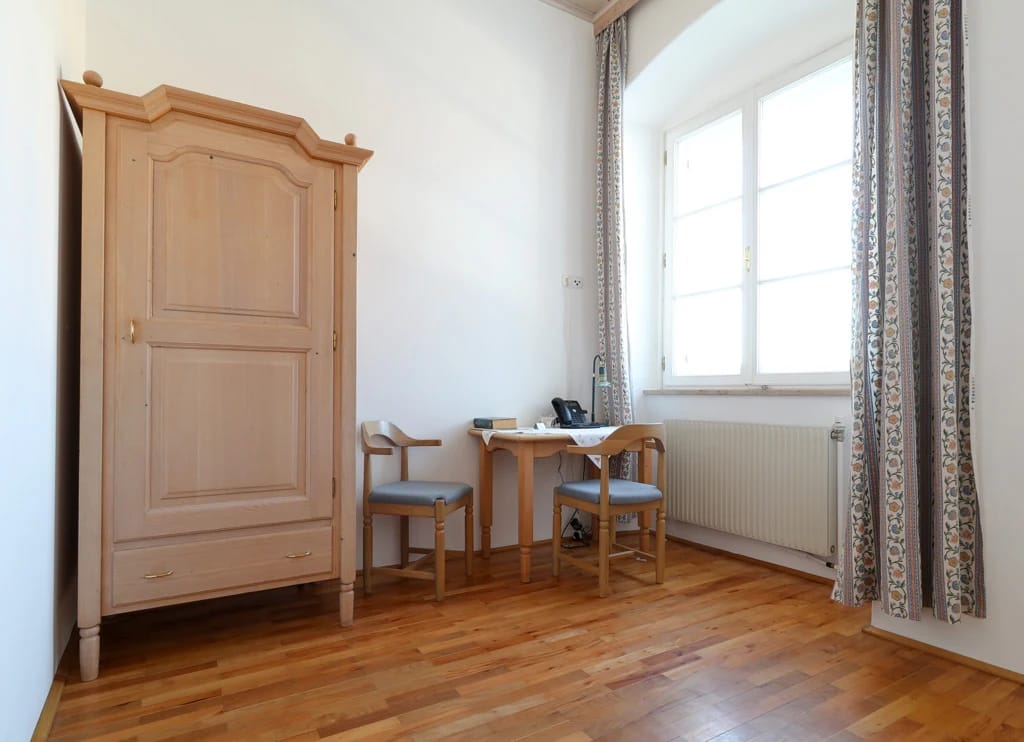 Zimmer mit hölzernem Kleiderschrank, kleinem Schreibtisch und zwei Stühlen am Fenster