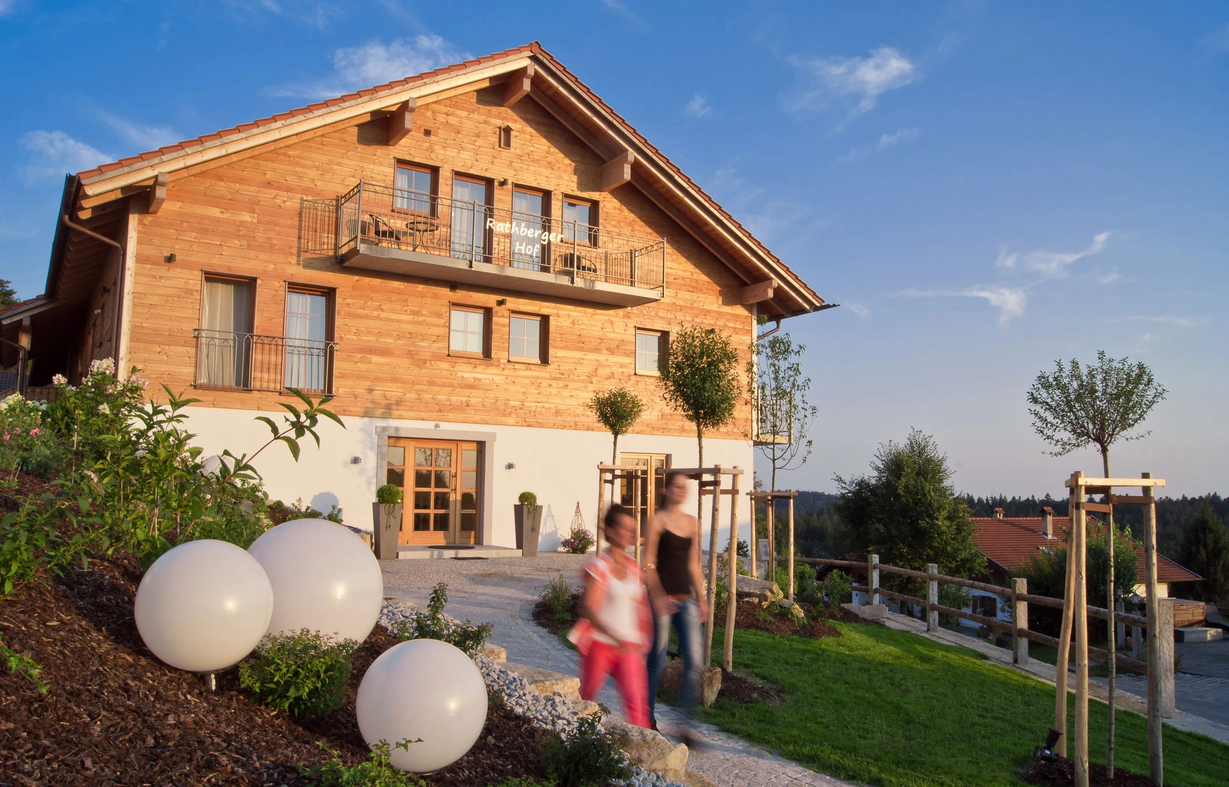 Holzverkleidetes Haupthaus mit Balkonen und Eingang, davor Gartenweg mit Kugelleuchten und jungen Bäumen im Abendlicht