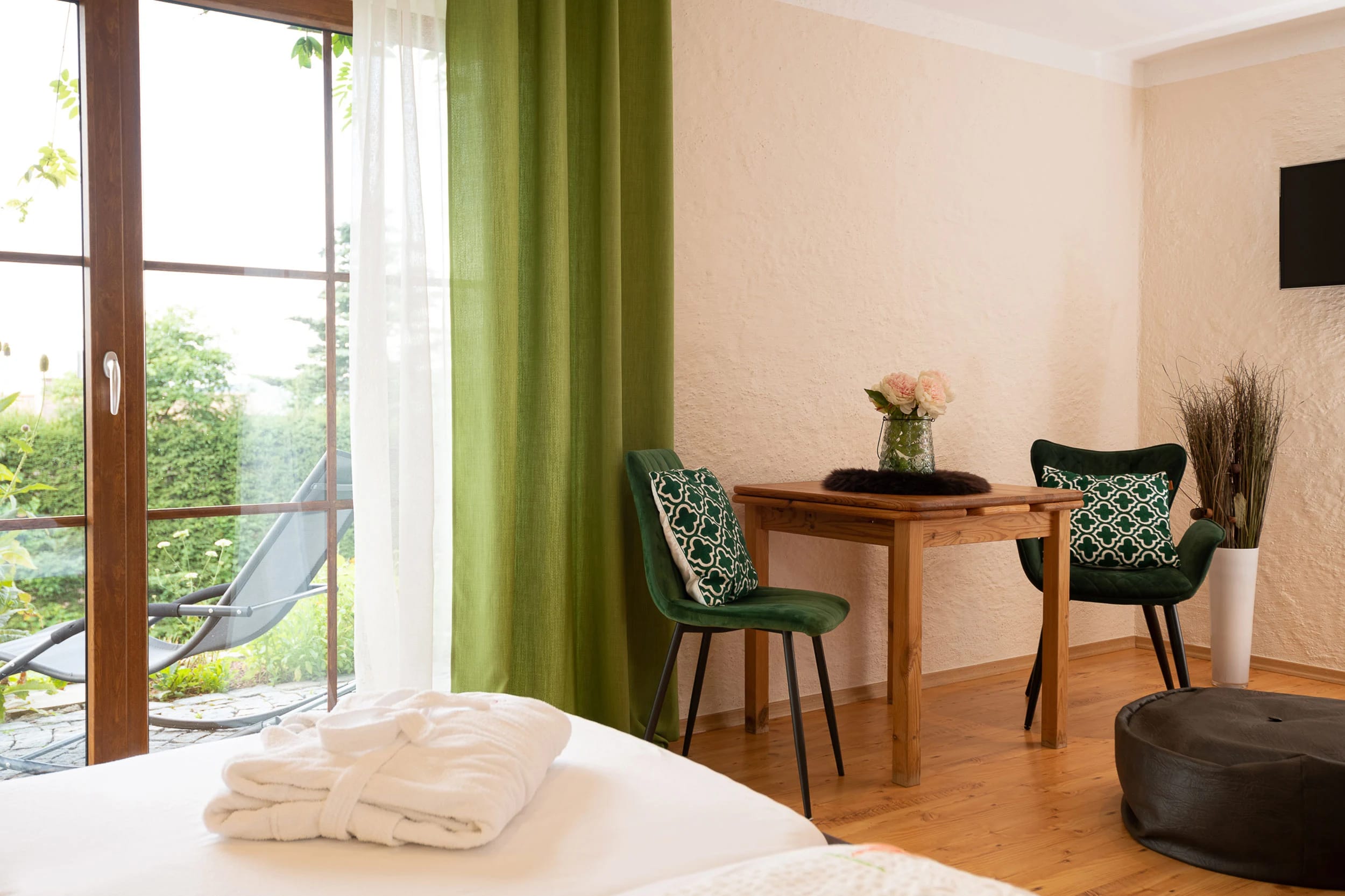 Zimmer mit Bademänteln auf dem Bett, grünem Vorhang, Balkontür und Schreibtisch mit zwei grünen Sesseln