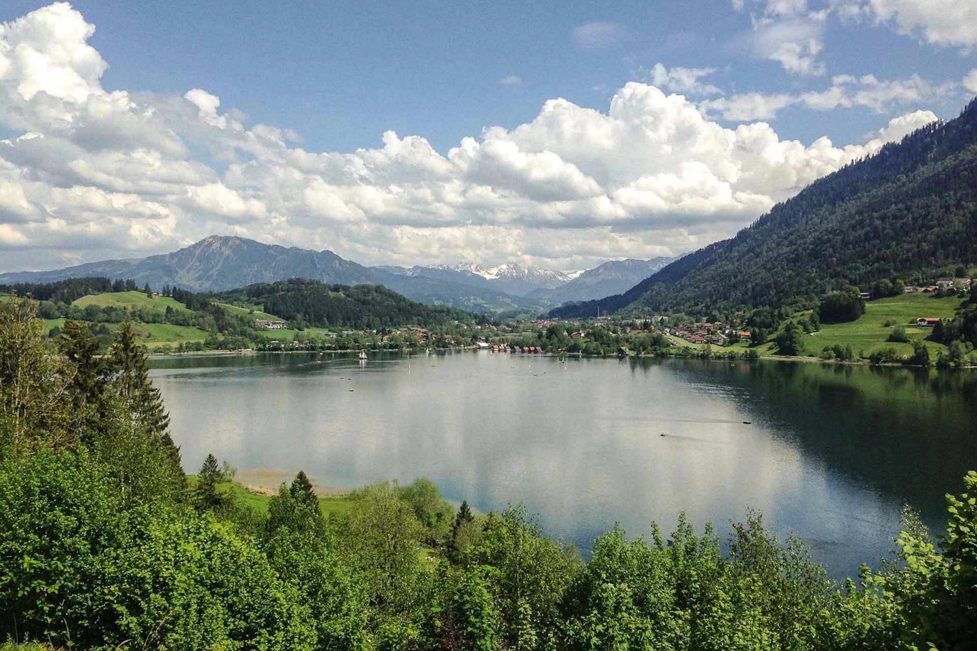 Blick über den Alpsee mit bewaldeten Ufern und Bergen unter Wolkenhimmel