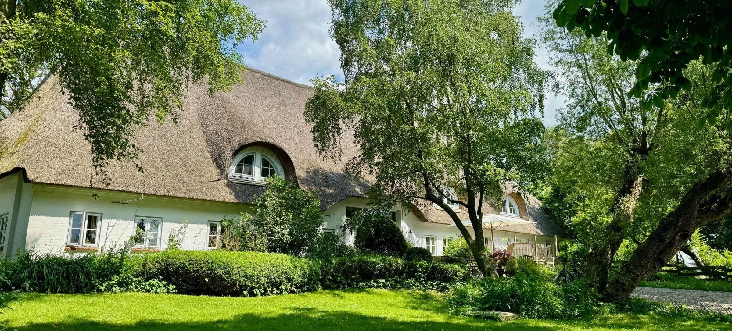 Reetgedecktes Haupthaus mit Rasenfläche, Hecken und alten Bäumen im Sommer