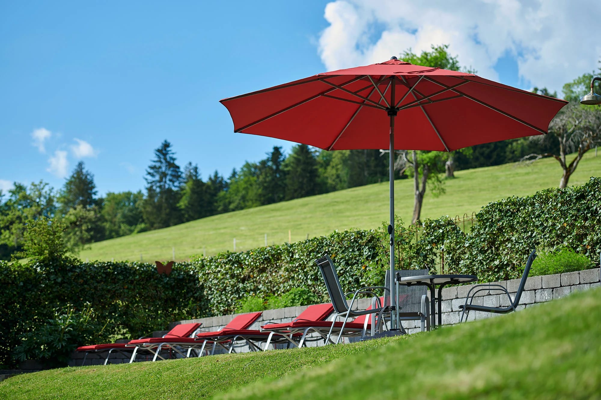 Sonnenterrasse mit Liegestühlen unter rotem Sonnenschirm, dahinter Hecke und grüne Wiese