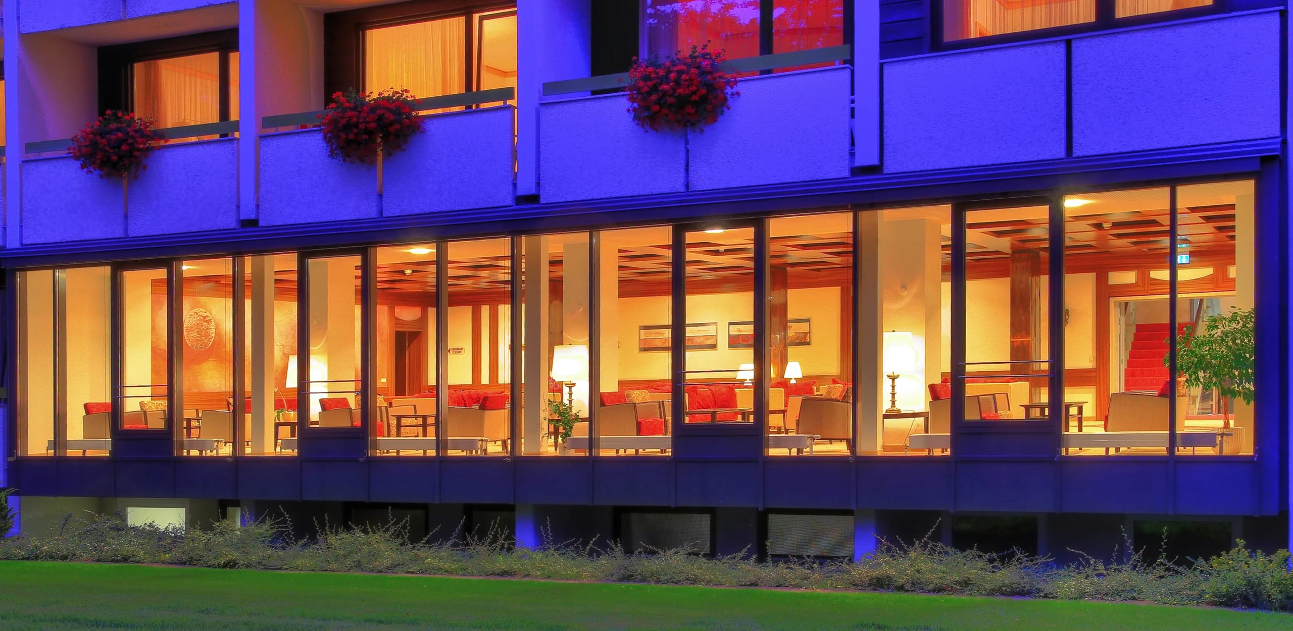 Beleuchtete Fensterfront des Aufenthaltsbereichs am Abend, mit Blumenkästen an den Balkonen