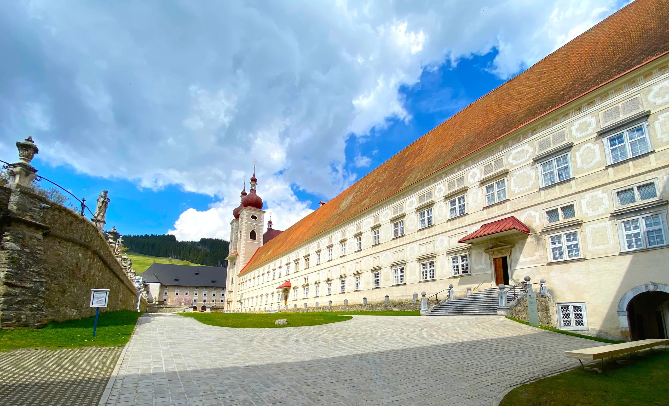 Stiftshof mit langem Klostergebäude, Kirchturm und Steinbalustrade unter blauem Himmel
