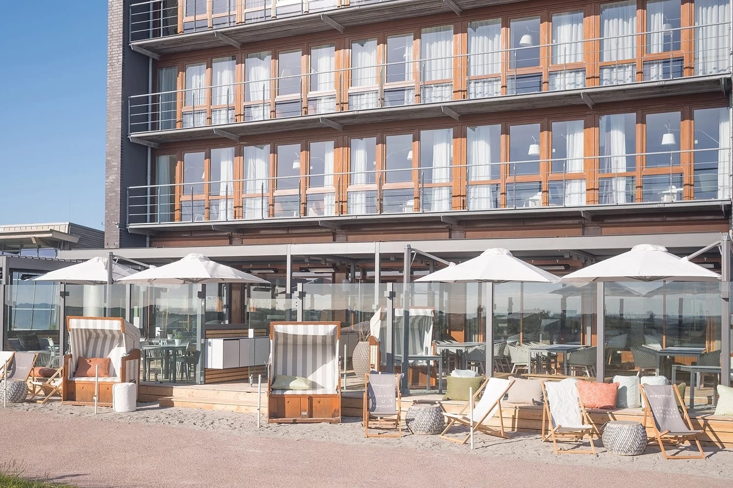 Außenansicht des Resorts mit Holzfassade und Balkonen, davor die Terrasse mit Sonnenschirmen, Strandkörben und Liegestühlen
