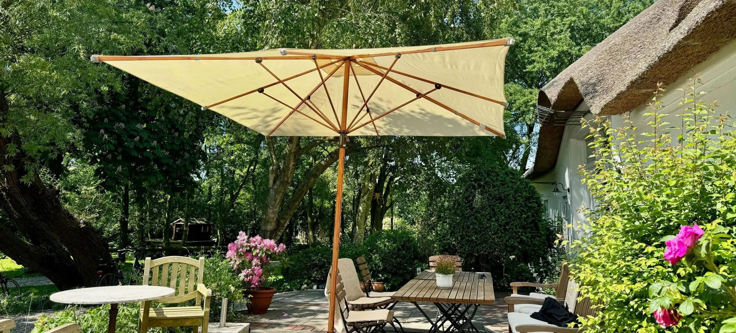 Terrasse mit großem Sonnenschirm, Holztisch, Stühlen und Blumenkübeln neben dem reetgedeckten Haus