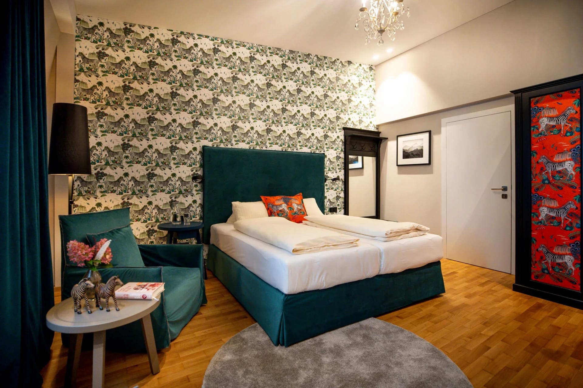 Zimmer mit grün gemusterter Tapete, türkisfarbenem Bett, Sessel und rot gemustertem Schrank