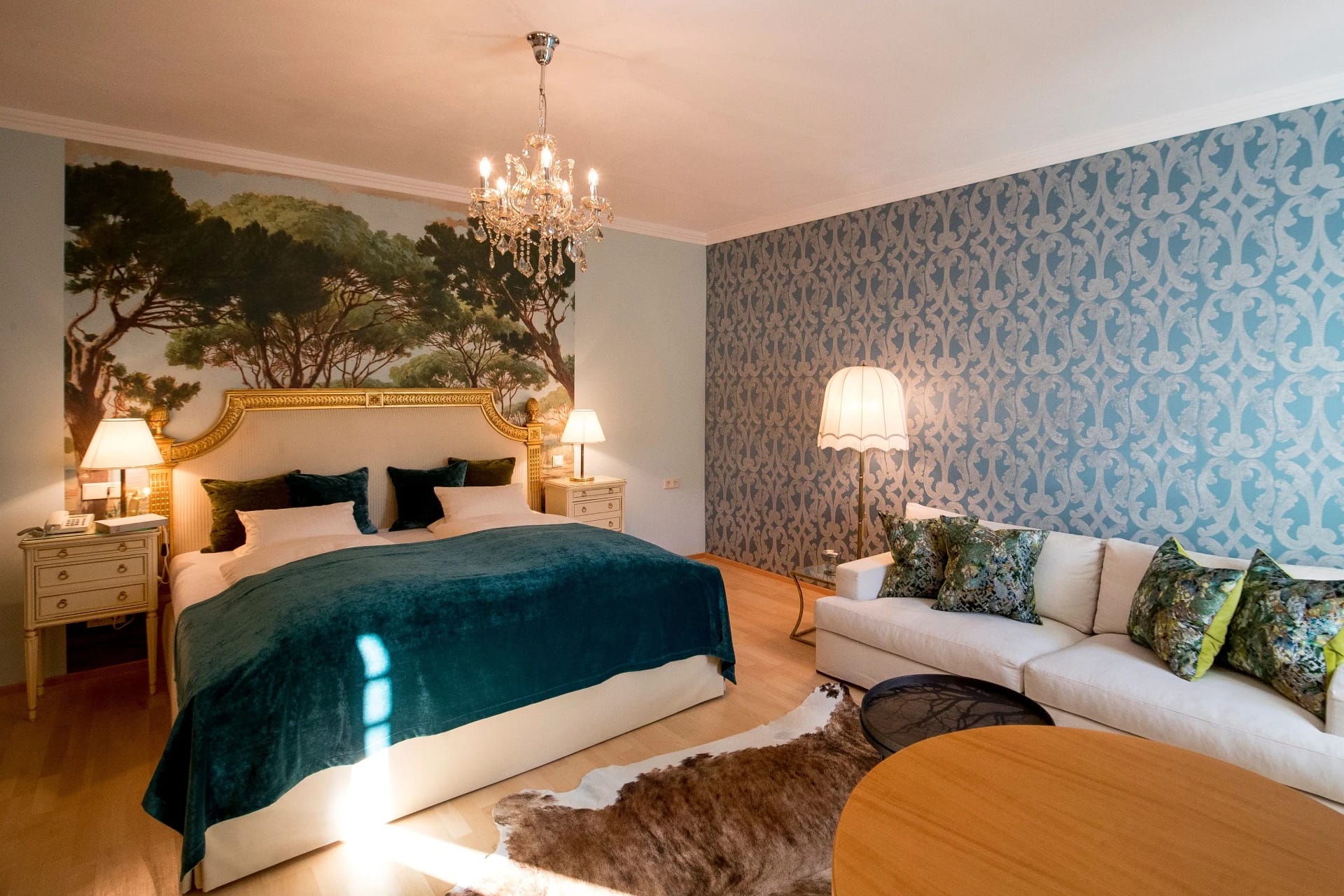 Zimmer mit türkisfarbenem Bett vor gemaltem Wandbild, Kronleuchter, heller Couch und Fellteppich