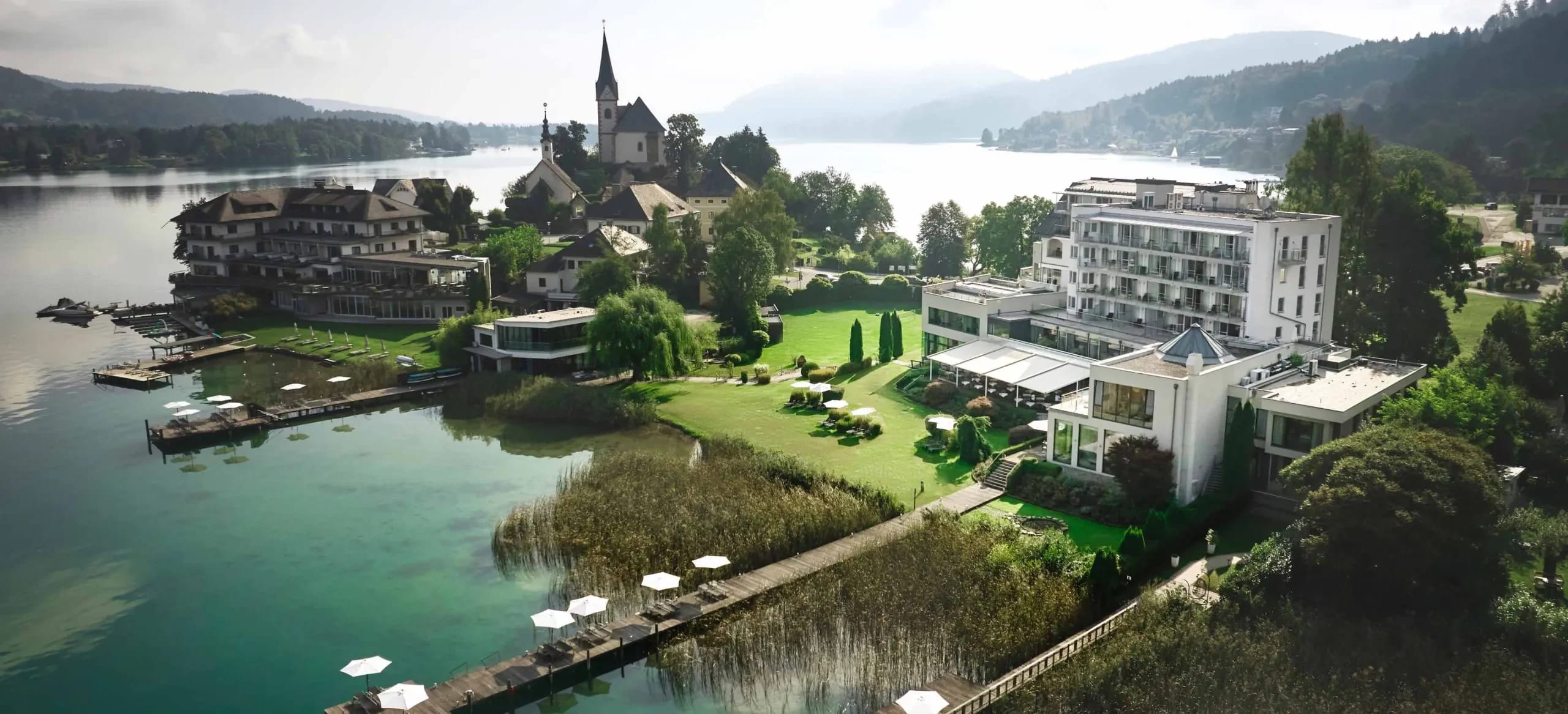 Luftaufnahme des Hotelgebäudes und der Gartenanlage am Seeufer, links die Halbinsel mit der Kirche von Maria Wörth