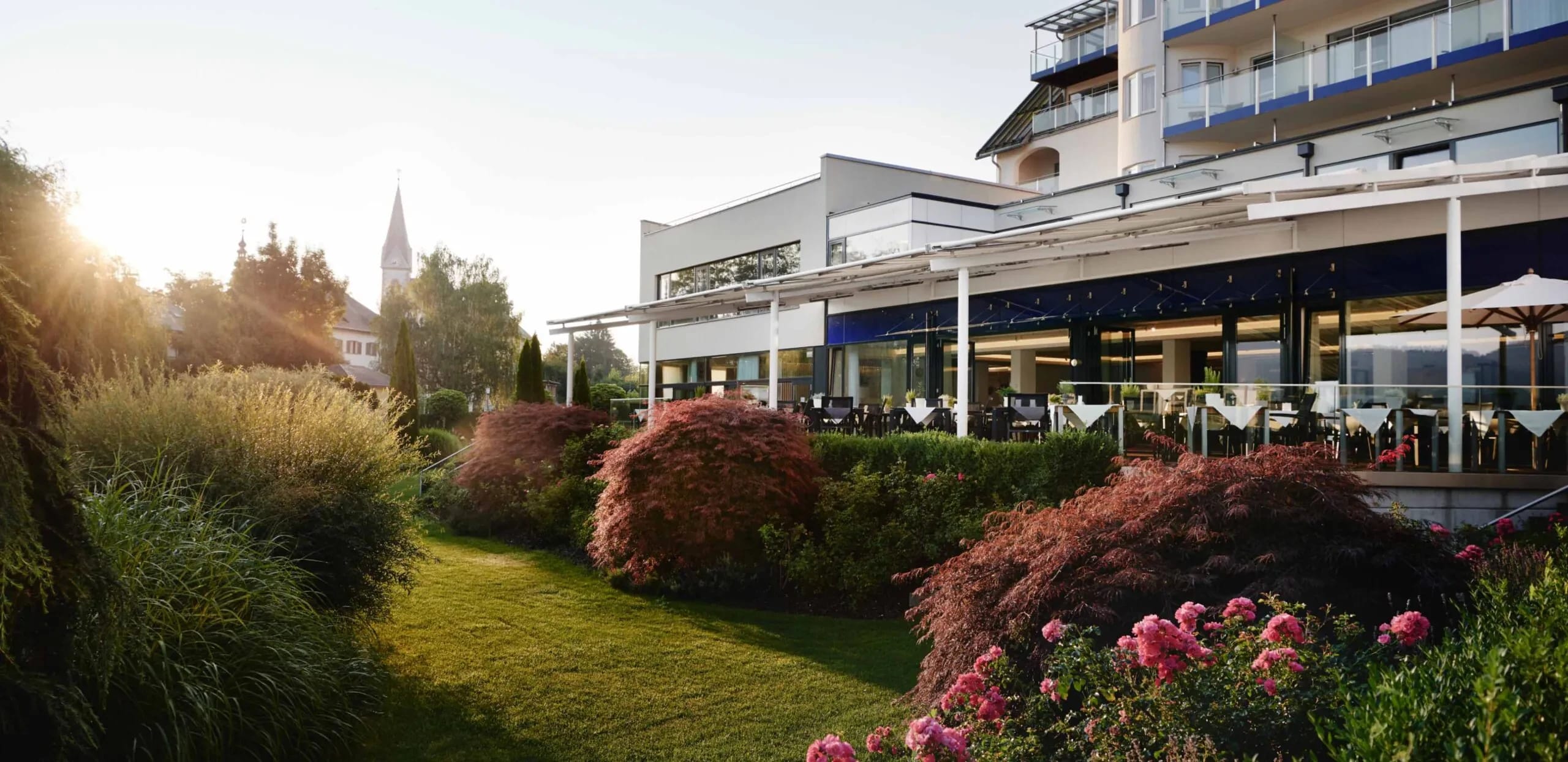 Garten mit blühenden Sträuchern und Rasen vor der überdachten Terrasse des Hauses in der Abendsonne