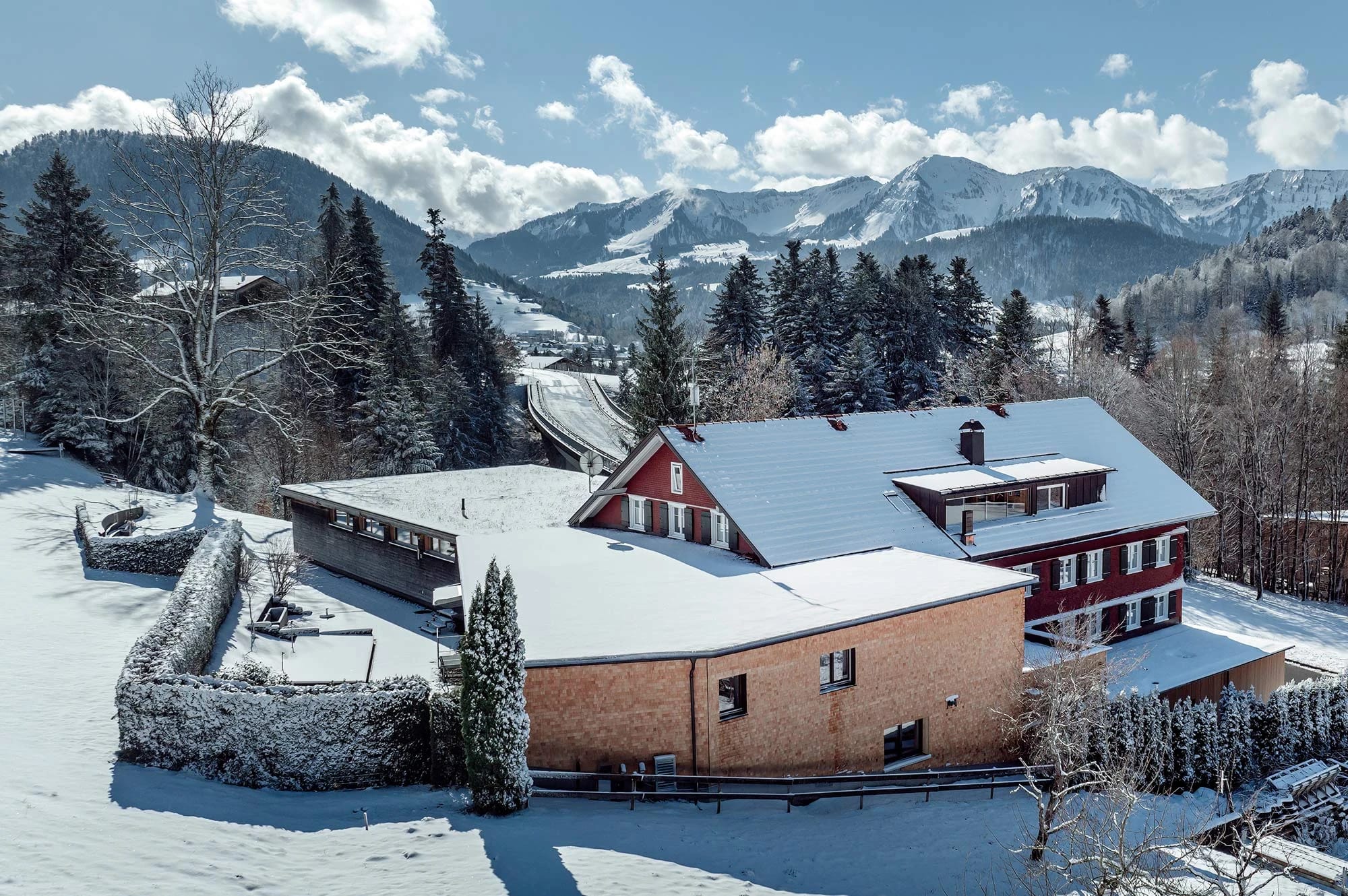 Verschneites Haus mit Nebengebäuden von oben, dahinter Wald und Bergkette im Winter