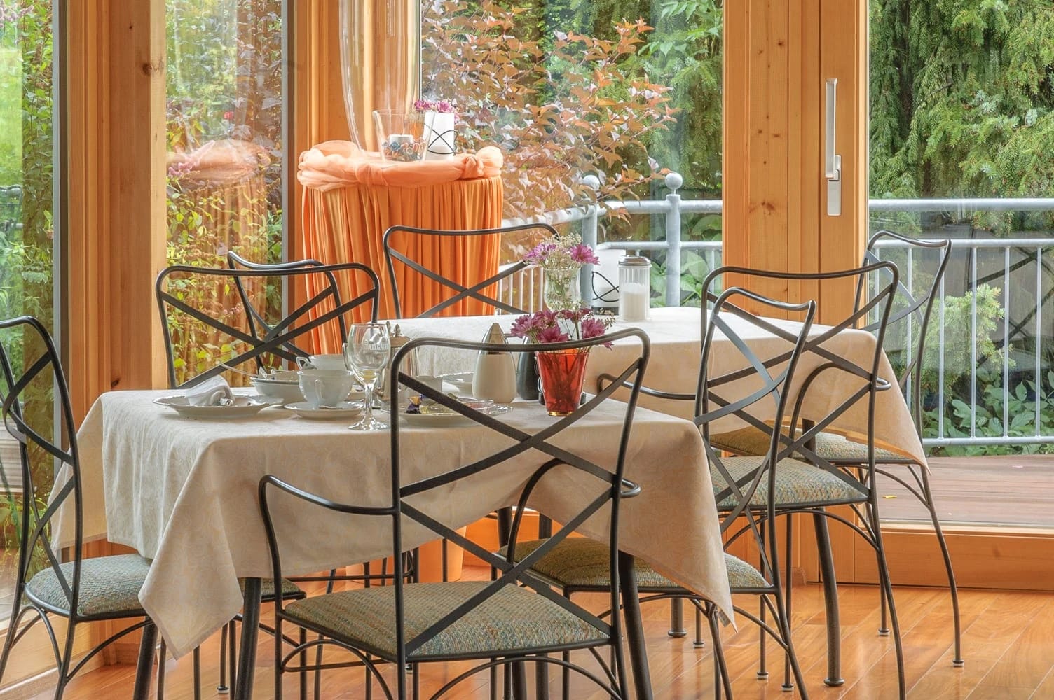 Wintergarten mit eingedeckten Tischen, Schmiedeeisenstühlen und Fensterfront ins Grüne