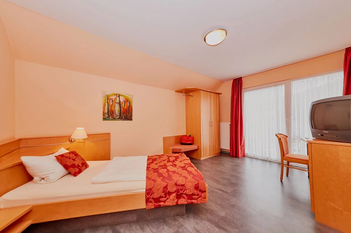 Zimmer mit Doppelbett, Kleiderschrank und Balkontür mit roten Vorhängen